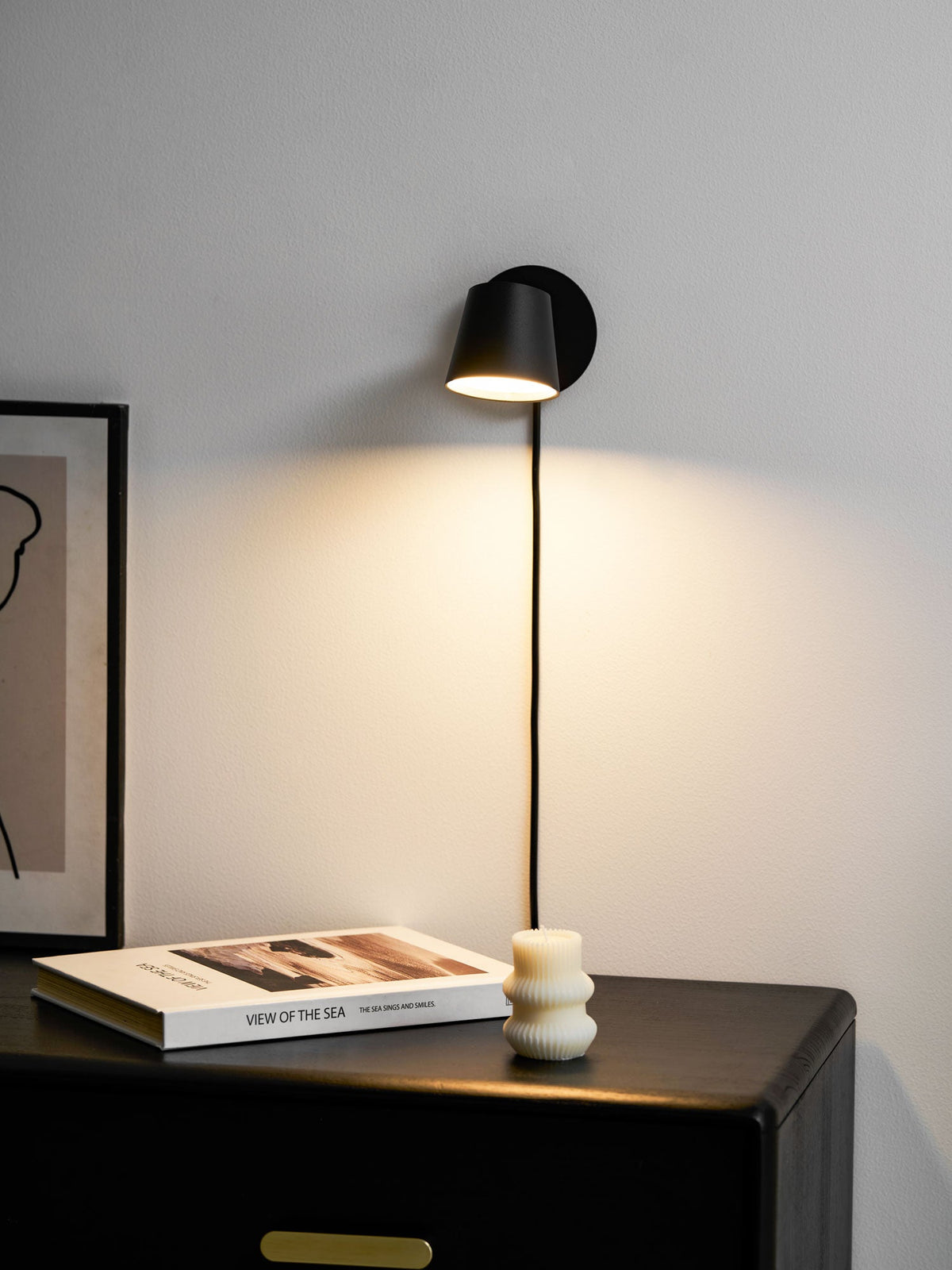 Warm light from a black Clérachic wall light falling across a white wall above a book, a ribbed candle and a framed print.