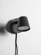 Close view of the black Clérachic wall light with its shade tilted forward and its cord hanging from the round backplate.