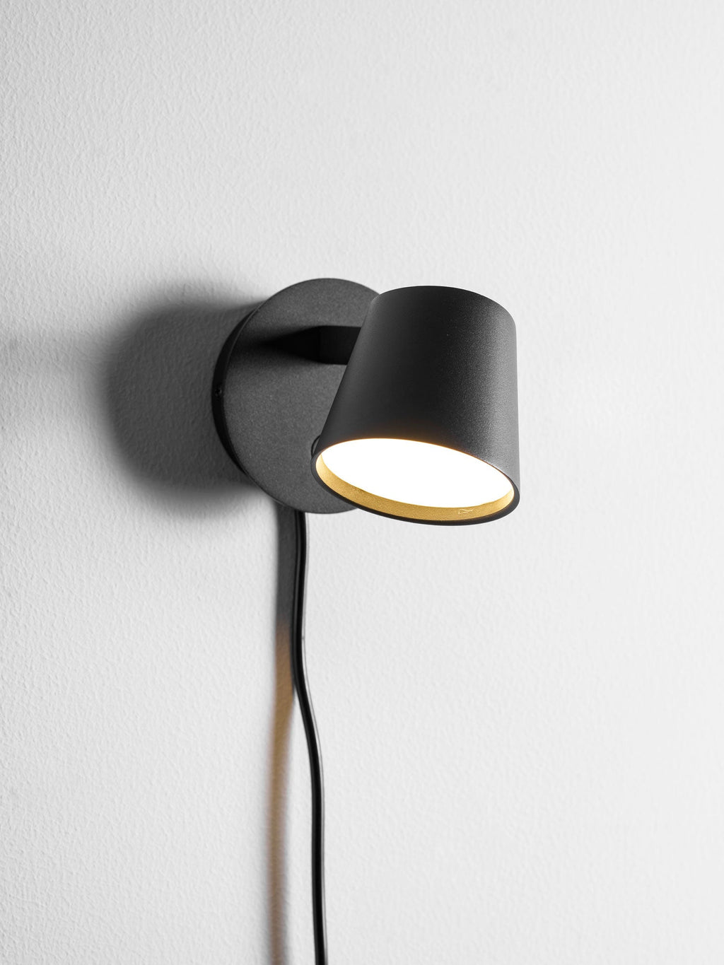 Close view of the lit black Clérachic wall light, its warm inner glow showing under the tilted shade on a white wall.