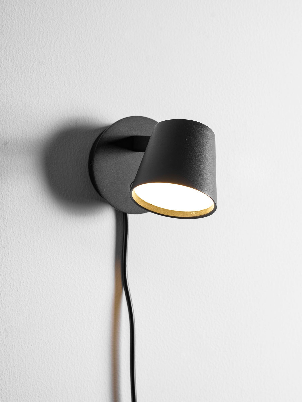 Close view of the lit black Clérachic wall light, its warm inner glow showing under the tilted shade on a white wall.