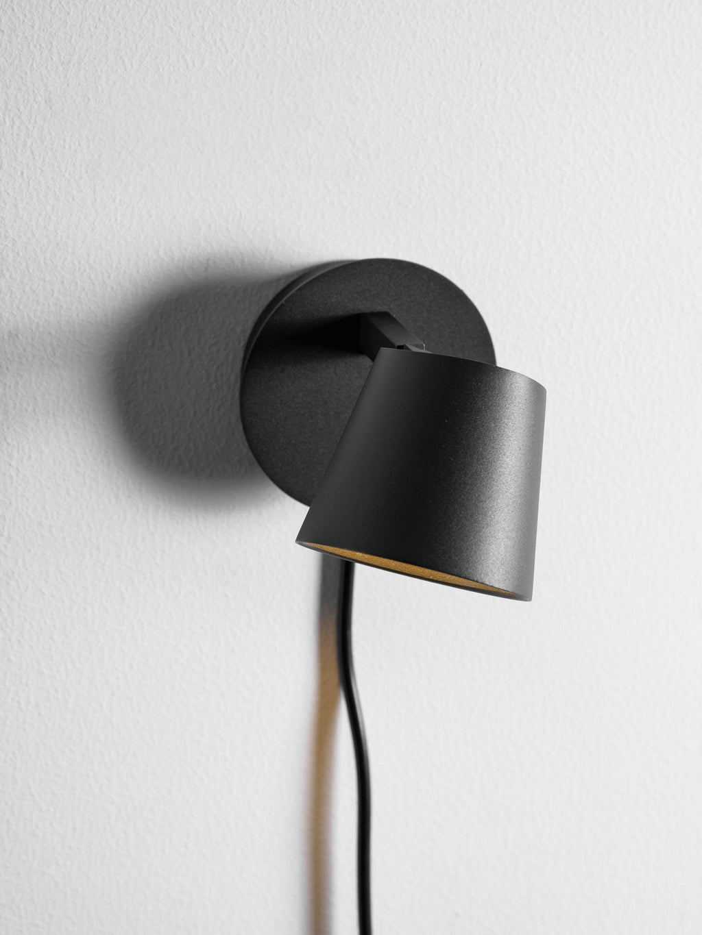 Black Clérachic wall light with the shade swung low in front of the backplate, close up on a white wall.