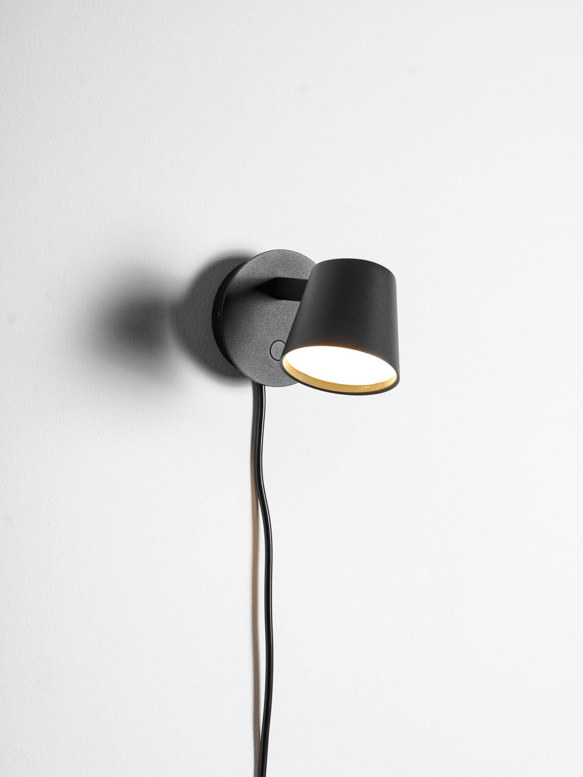 Black Clérachic wall light lit and seen close up, with a button on the backplate and the cord hanging below.
