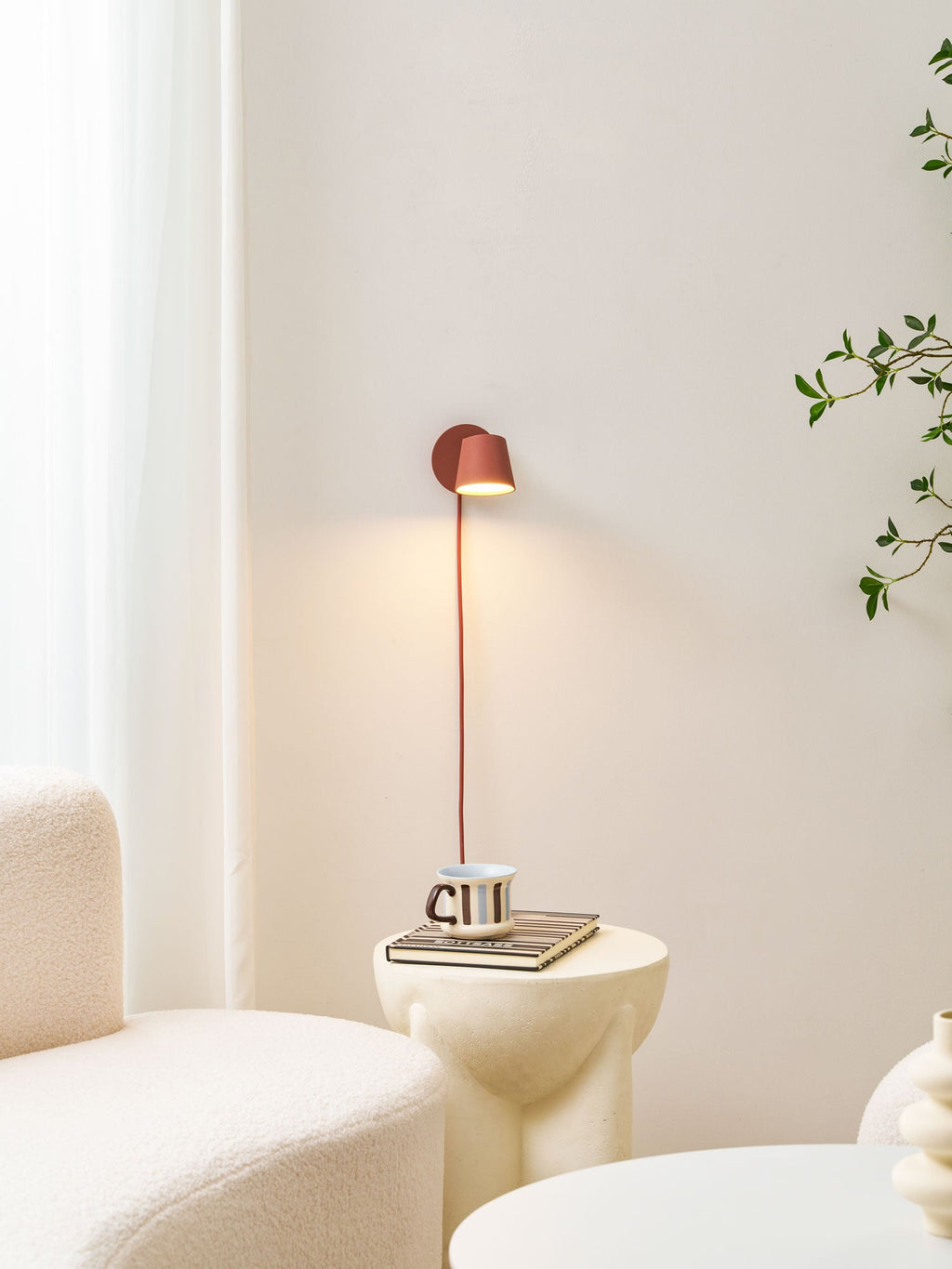 Small lit red Clérachic wall light on a cream wall above a ribbed white side table with a mug and boucle seating.