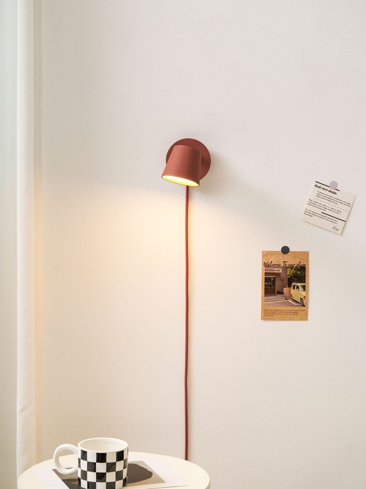 Lit red Clérachic wall light on a cream wall, a red cord below it and a checkered mug on the side table.
