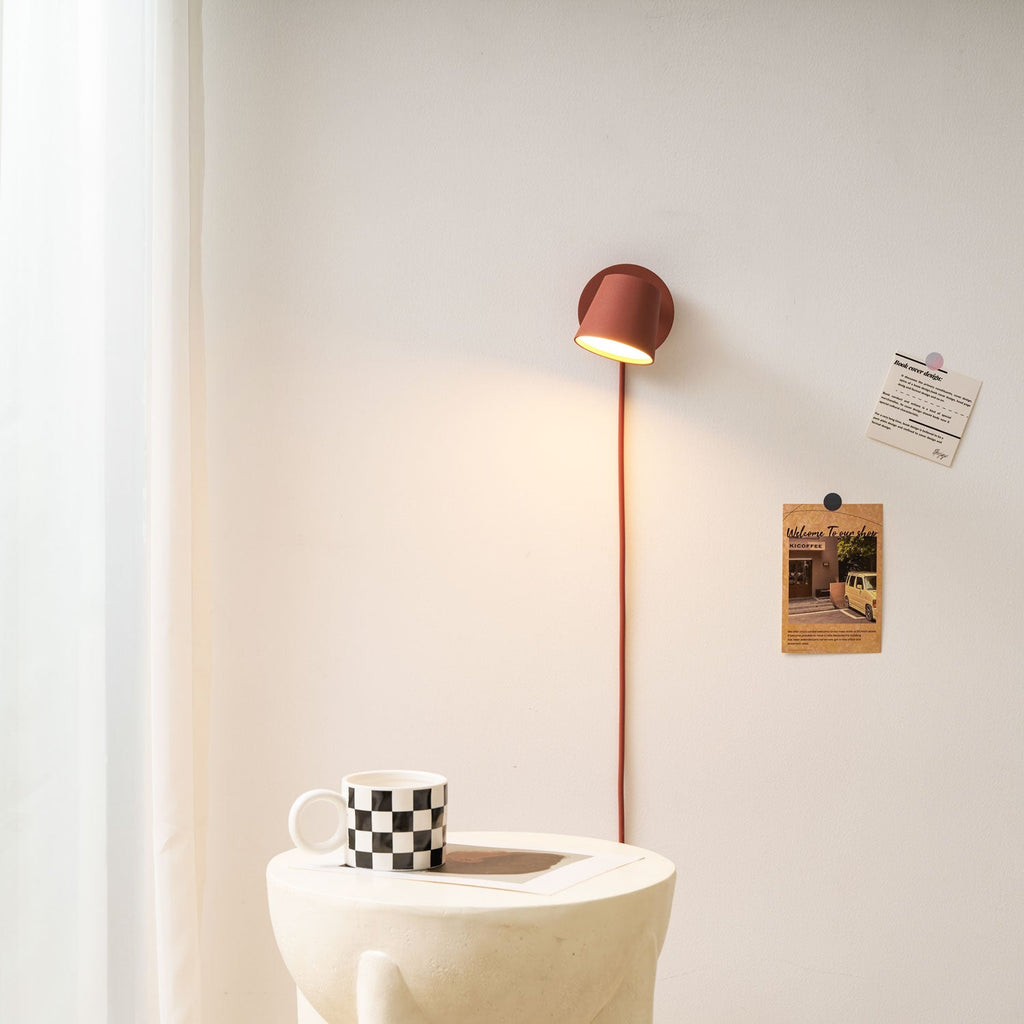 Small red Clérachic wall light glowing above a round white side table with a checkered mug and curtains at the left.