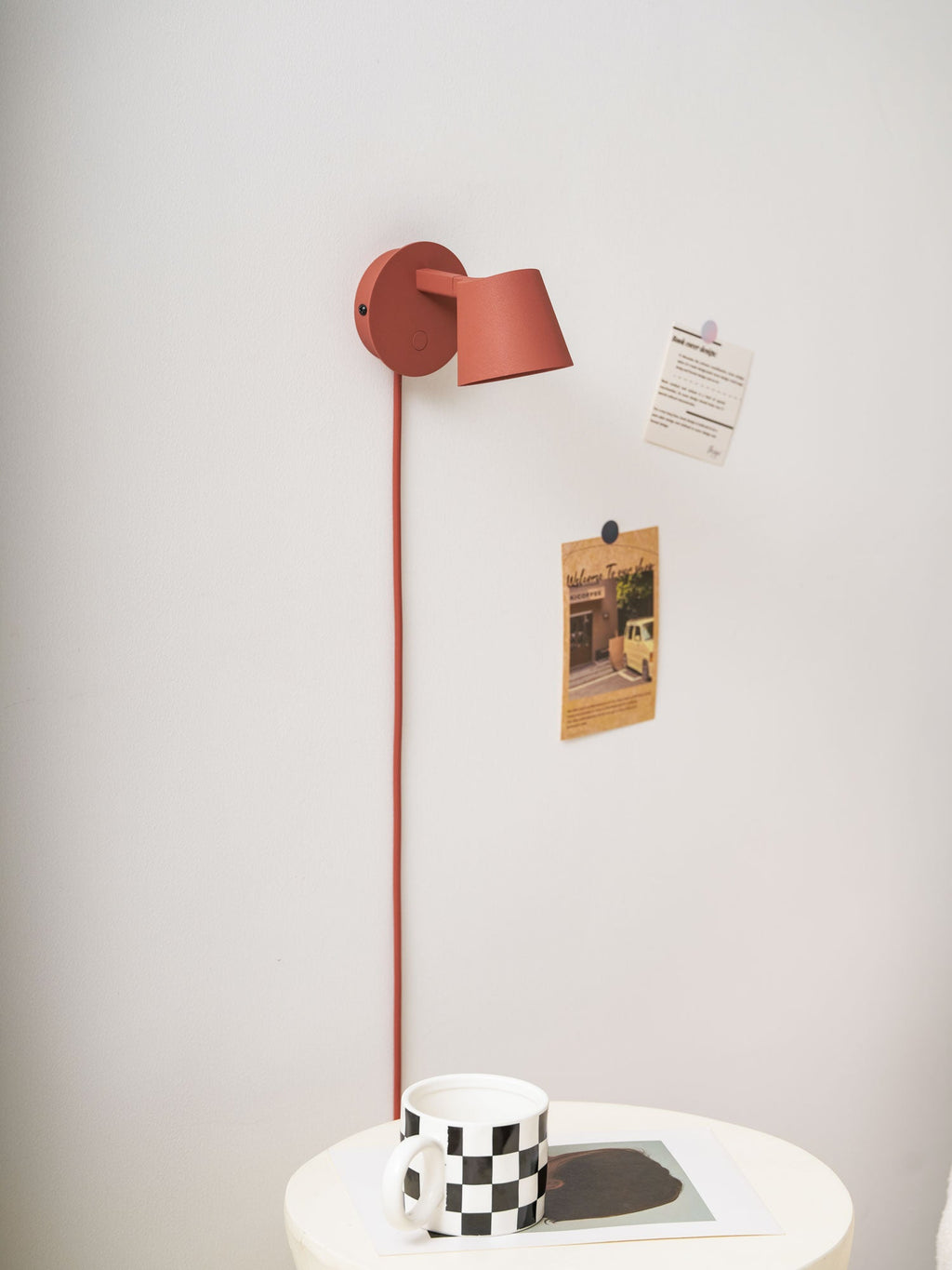 Red Clérachic wall light turned side-on above a checkered mug, with pinned notes on a pale wall.