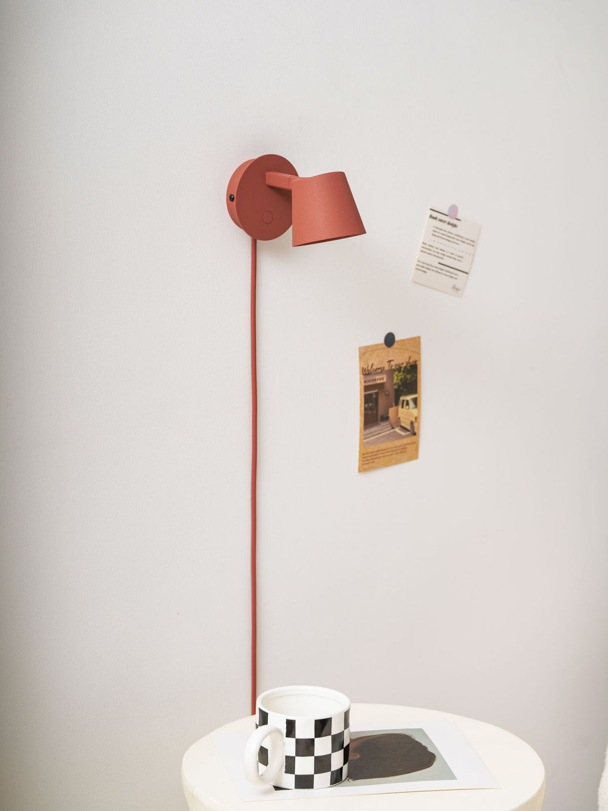 Red Clérachic wall light turned side-on above a checkered mug, with pinned notes on a pale wall.