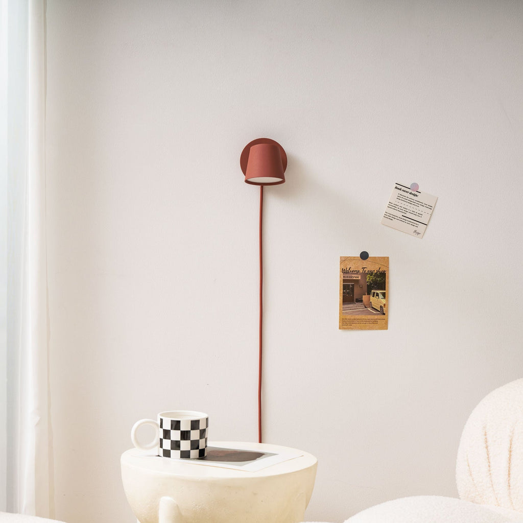 Red Clérachic wall light with its shade facing down, its cord dropping to a checkered mug on a cream side table.