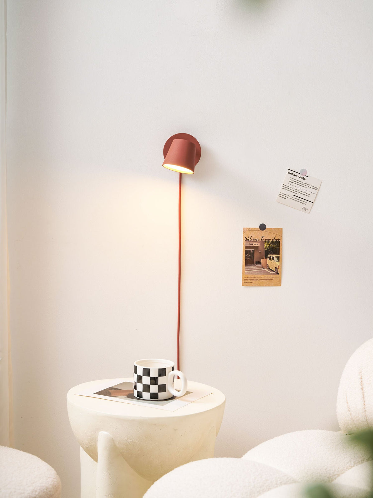 Lit red Clérachic wall light above a checkered mug on a round table, with boucle seating and a cream wall behind.