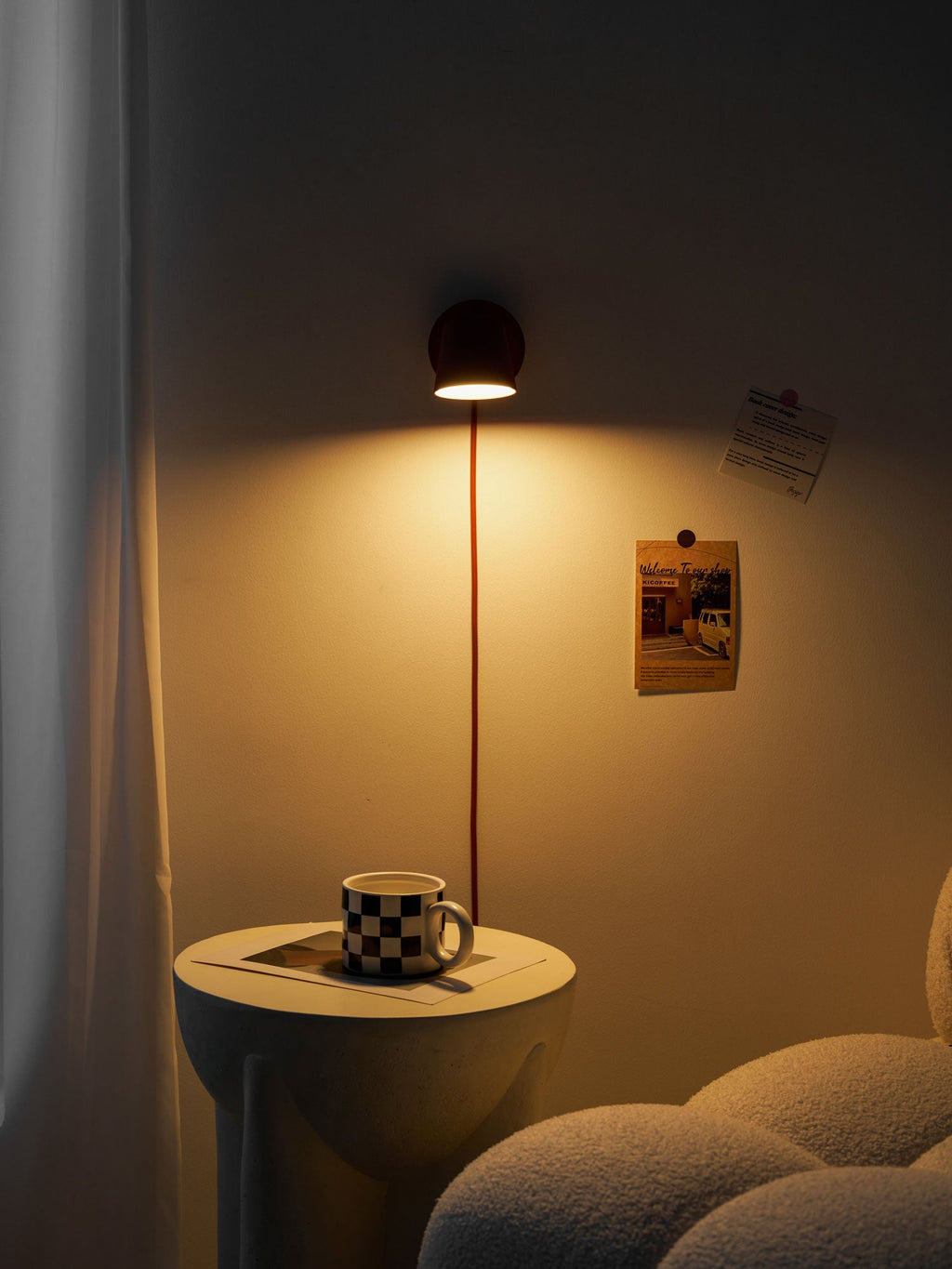 Red Clérachic wall light glowing in a dark room, warm light on the curtain and a mug on a side table below.