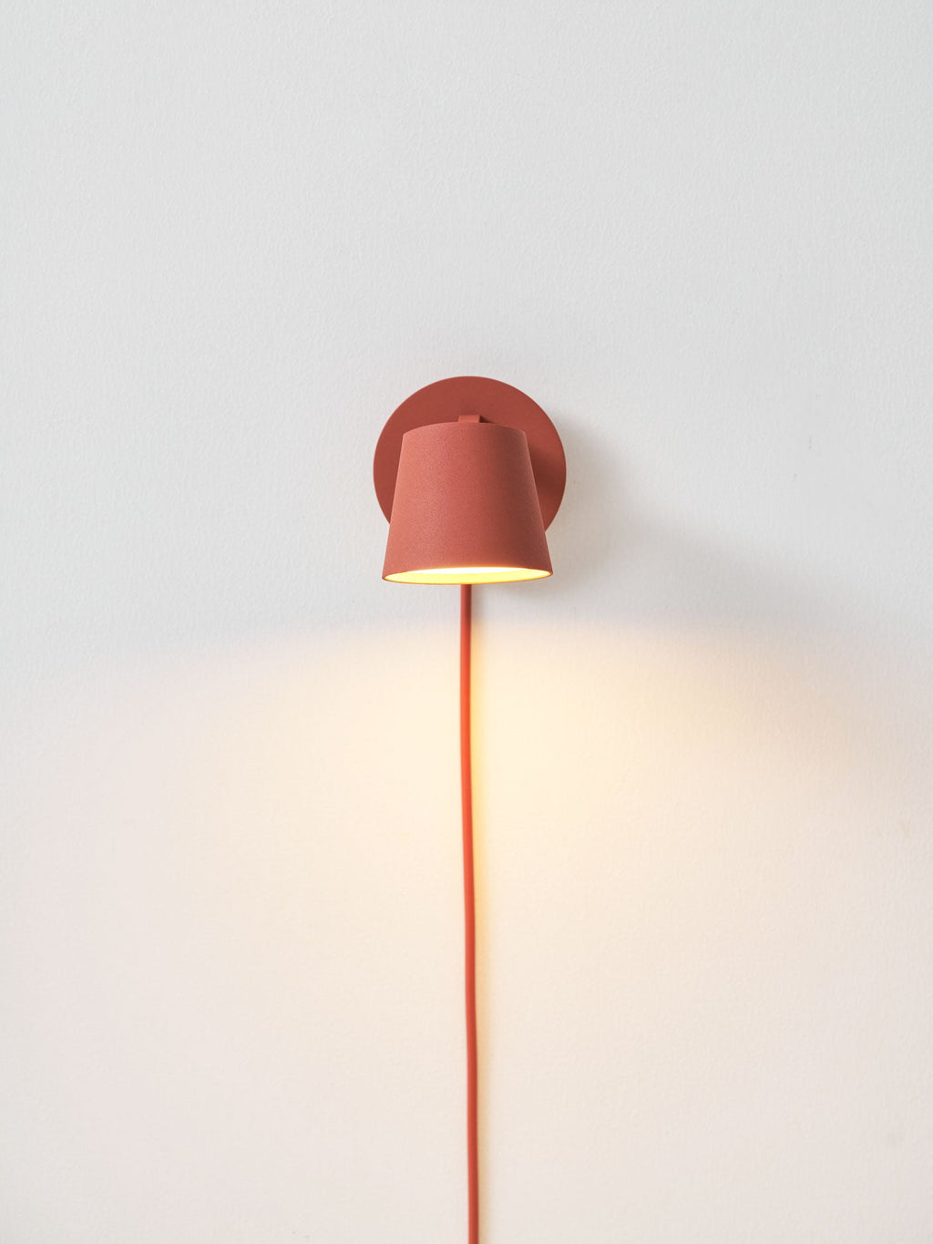 Small lit red Clérachic wall light on a pale wall with a warm wash of light below it and its red cord hanging down.