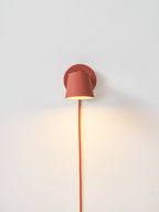 Small lit red Clérachic wall light on a pale wall with a warm wash of light below it and its red cord hanging down.