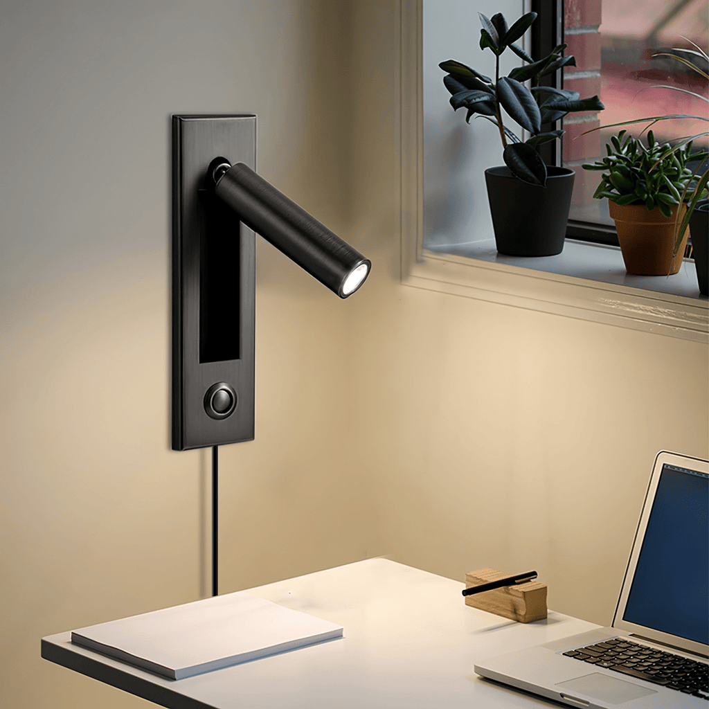 This Clérachic wall light in matt black has its round head angled out beside a window with plants, above a desk and laptop.