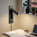This Clérachic wall light in matt black has its round head angled out beside a window with plants, above a desk and laptop.