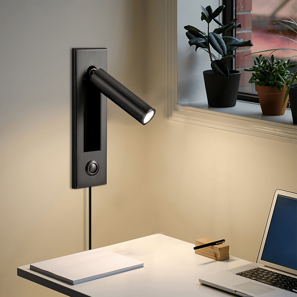 This Clérachic wall light in matt black has its round head angled out beside a window with plants, above a desk and laptop.