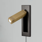 A Clérachic wall light with a dark grey plate and a brass-coloured square head angled out towards a pale pillow edge.