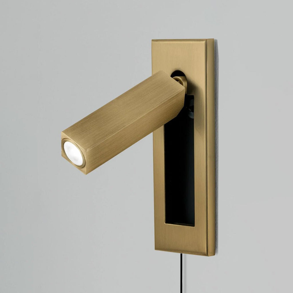 A brass-coloured plate and square head on this Clérachic wall light, with the LED lit and the cable hanging below.