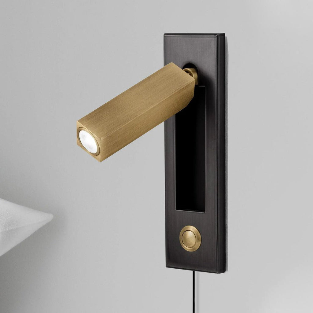 This Clérachic wall light has a dark plate, brass-coloured square head and a small round brass button on a grey wall.
