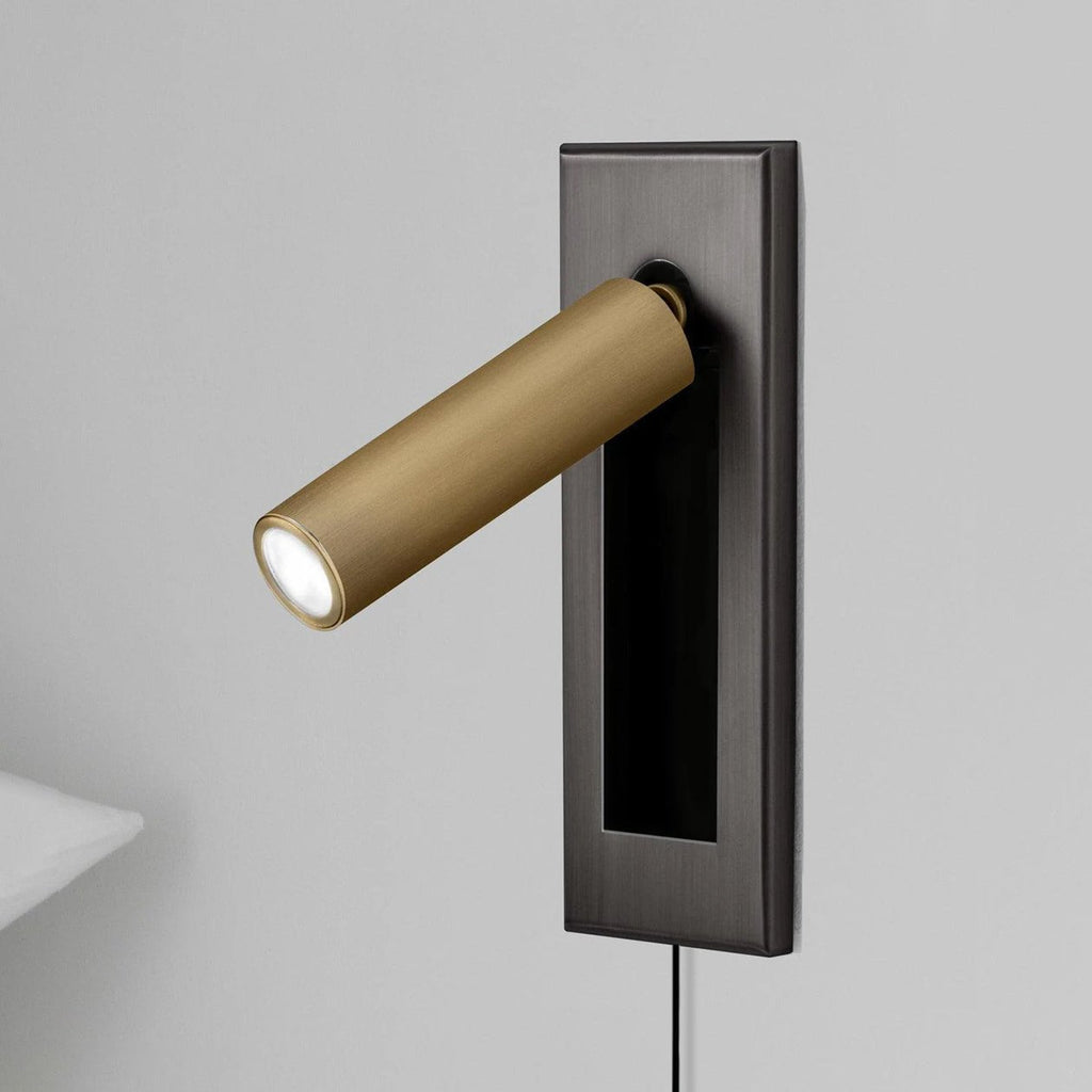 Round brass-coloured head of this Clérachic wall light angled out from a dark grey plate, cable trailing below.