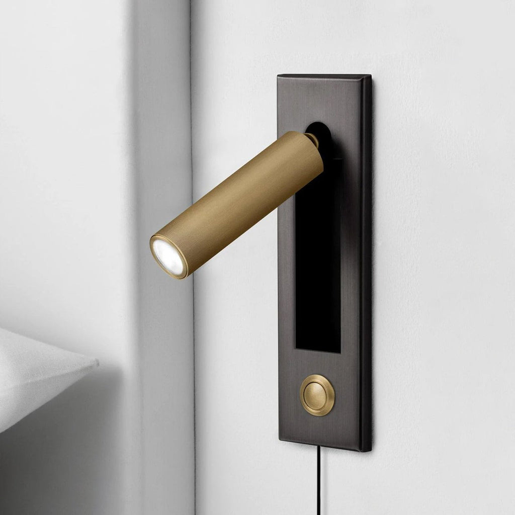 A Clérachic wall light on a dark plate with a round brass head and push button, set in a pale corner by a pillow.