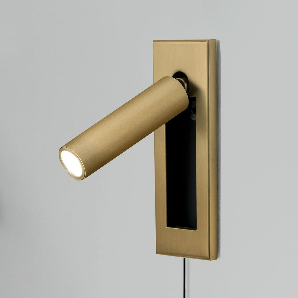 Round brass-coloured head and plate of this Clérachic wall light, lit and angled left, with a cable hanging down.