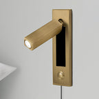 A square brass-coloured head and round button on a matching plate, shown on this Clérachic wall light against grey.