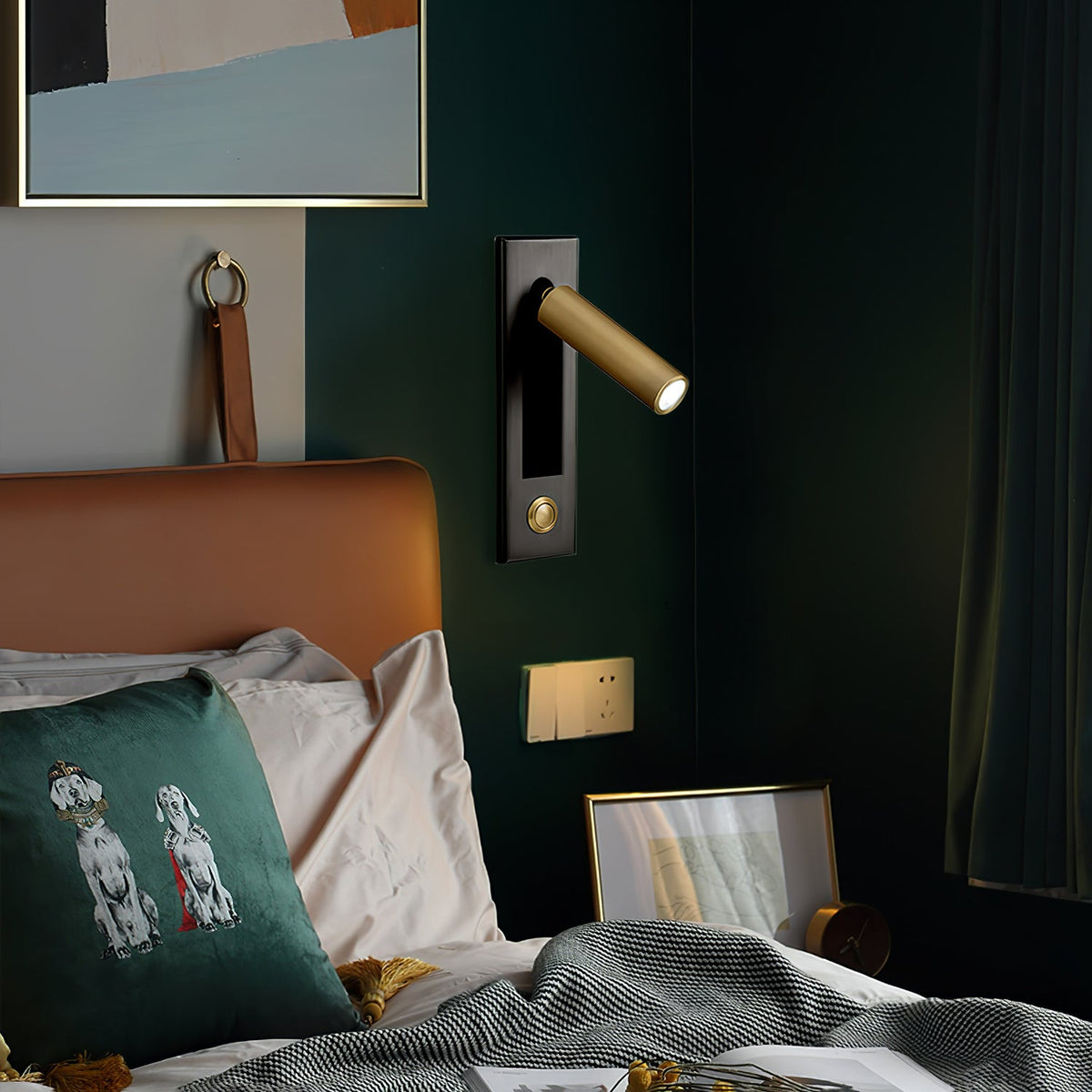 A Clérachic wall light with a slim brass-coloured head, lit above a headboard in a dark green bedroom with a cushion.