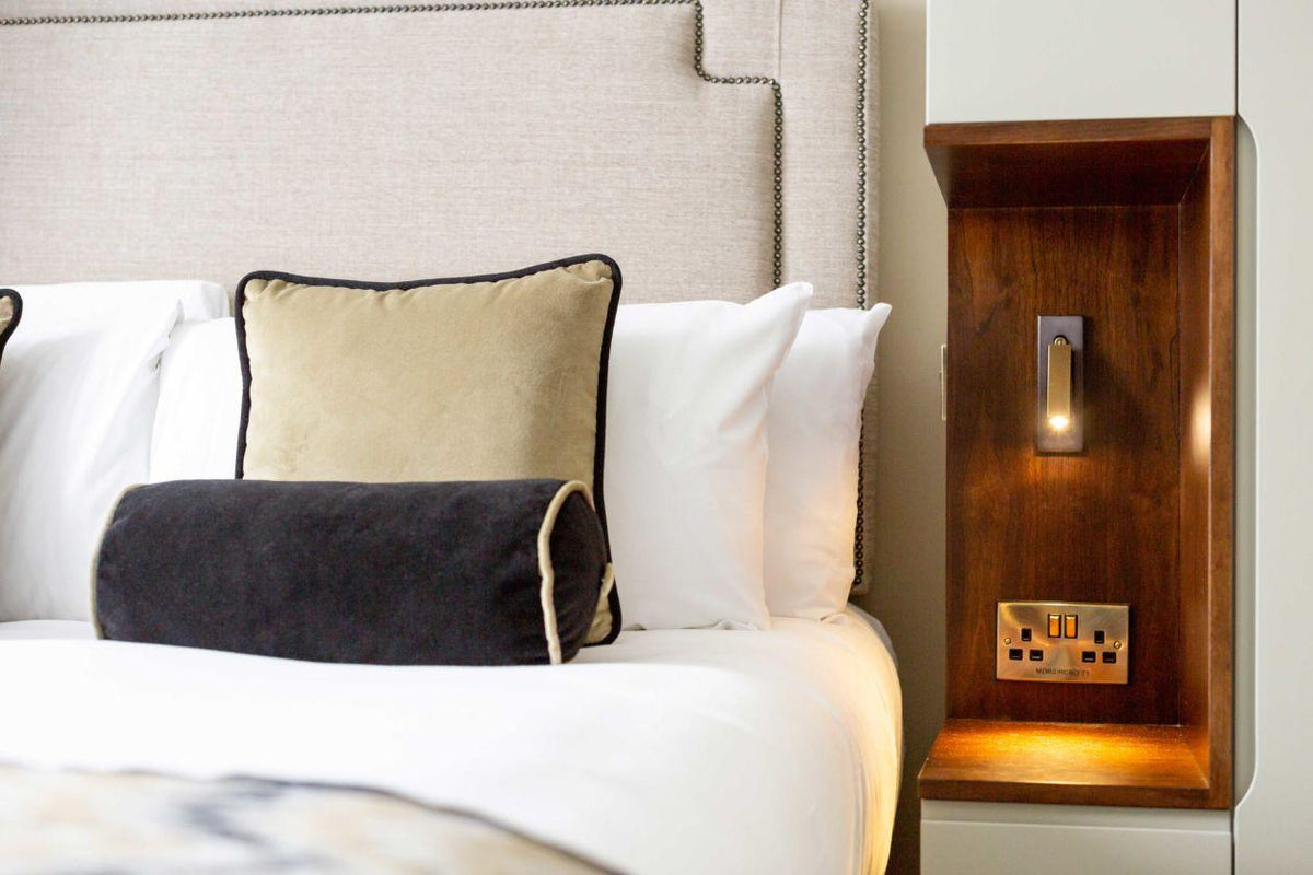 A Clérachic wall light glows in a wooden bedside niche above a brass socket, beside a bed with a headboard and cushions.