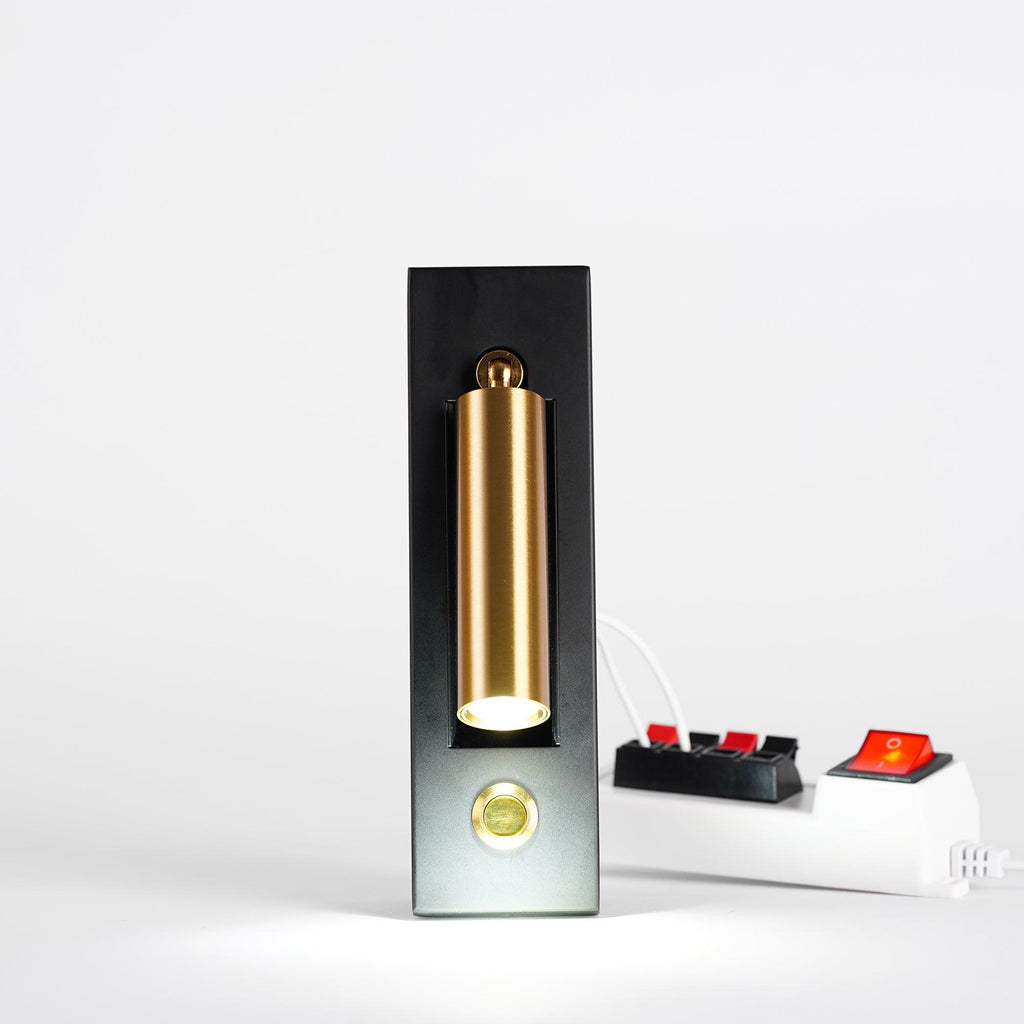 A Clérachic wall light standing on a plain surface with a brass-coloured head folded in, a lit LED, and a white power strip beside it.