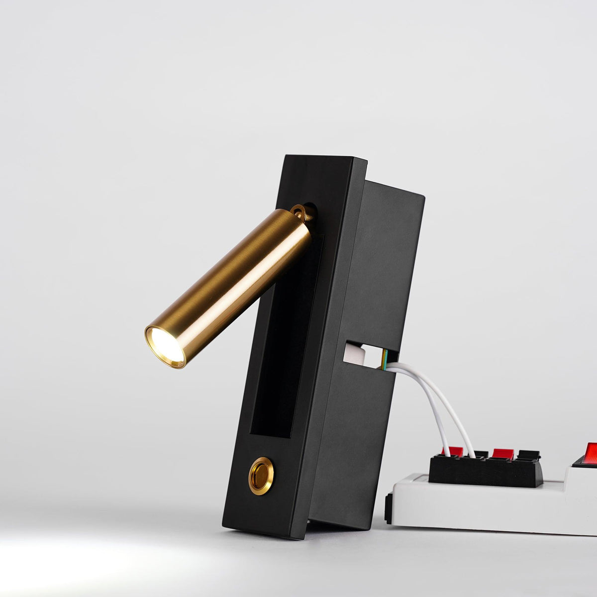 This Clérachic wall light stands on its black box with the brass-coloured head tilted out and lit, beside a white power strip.