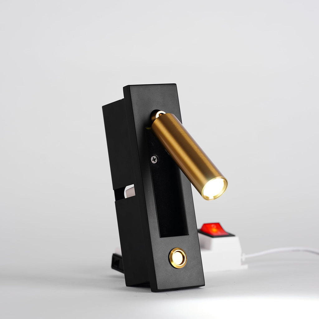 This Clérachic wall light on a surface shows the brass-coloured head lit and angled right, with a power strip behind it.