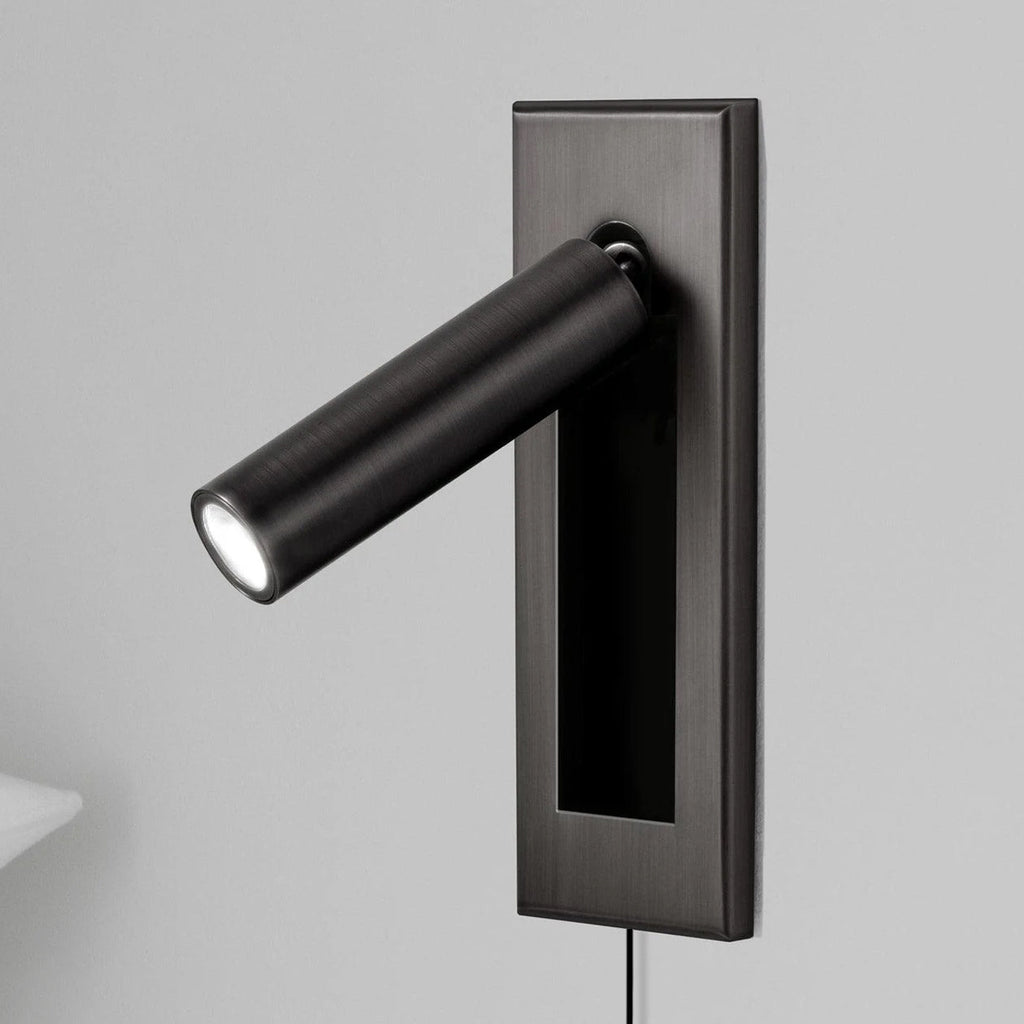 A Clérachic wall light with a dark plate and black round head, angled out and lit, with a pillow edge at the lower left.