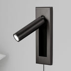 A Clérachic wall light with a dark plate and black round head, angled out and lit, with a pillow edge at the lower left.