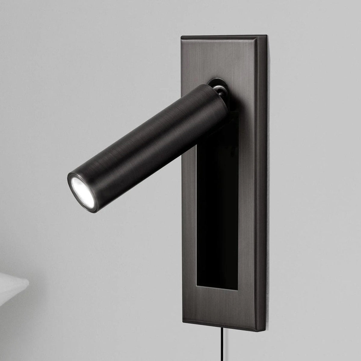 A Clérachic wall light with a dark plate and black round head, angled out and lit, with a pillow edge at the lower left.