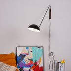Lit black Clérachic wall light reaching out from a walnut wall plate, above a colourful abstract canvas and an orange cushion.