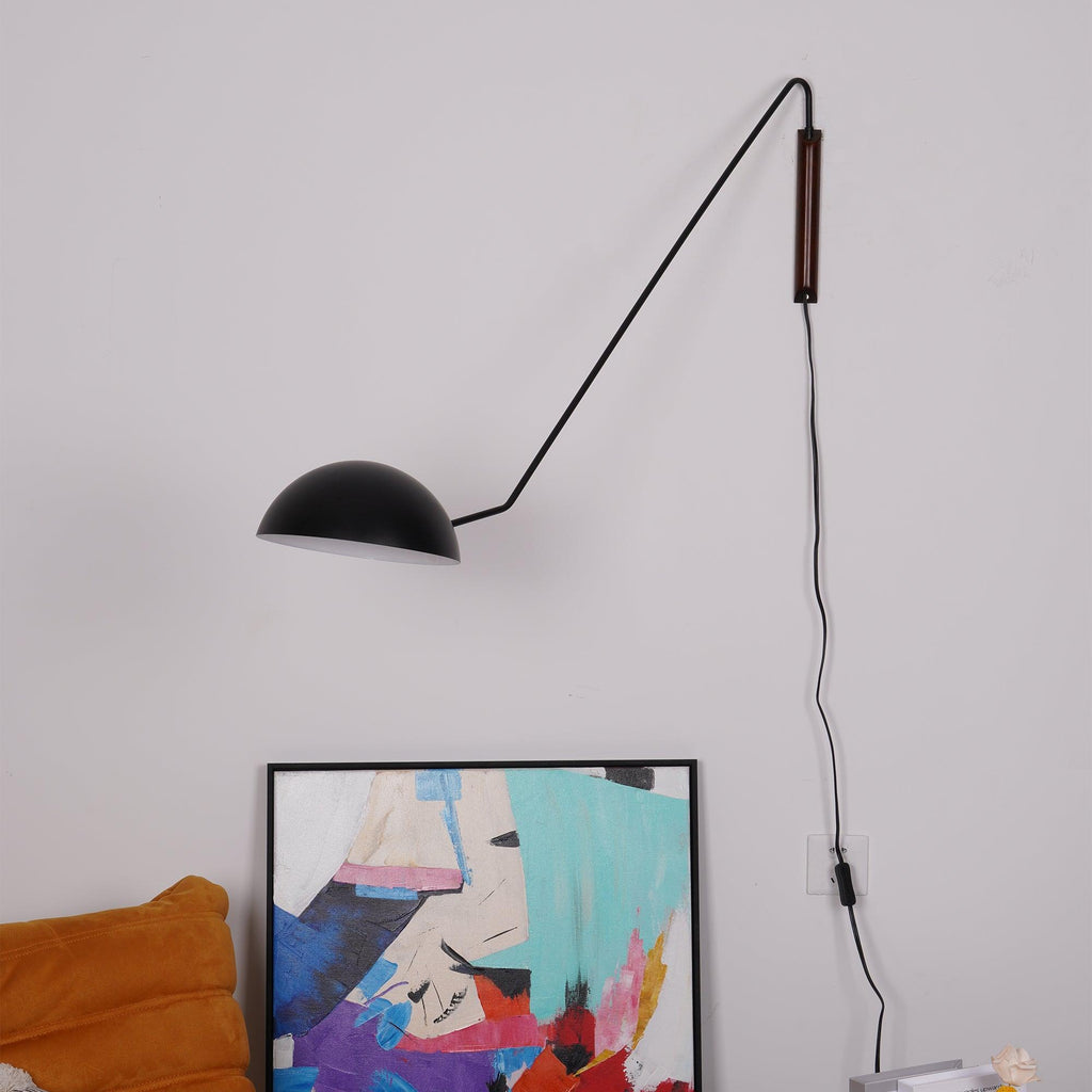 Unlit black Clérachic dome wall light on a long angled arm, with its cord hanging to a wall socket beside a framed abstract print.