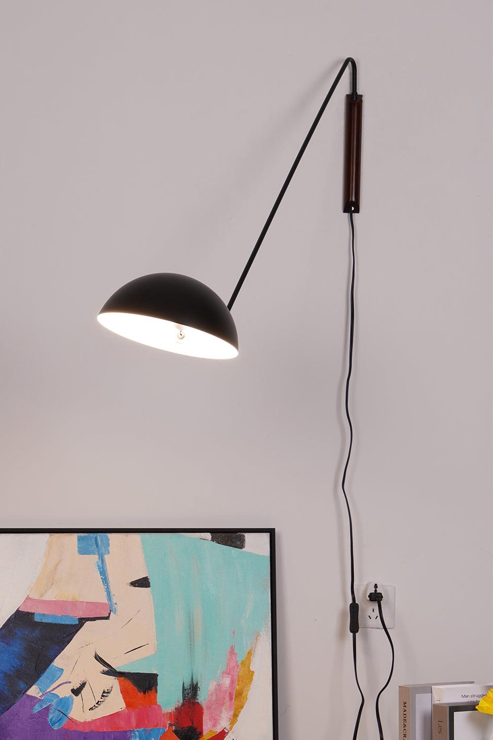 A black Clérachic wall light with a bright inner dome, its cord running down to a socket by a leaning abstract canvas.