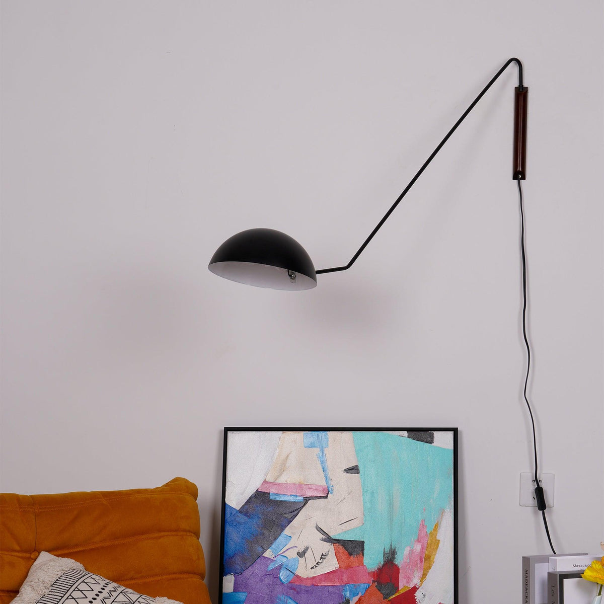 Black Clérachic swing-arm wall light seen from below the shade, over a leaning abstract painting and an orange cushion.