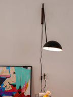 A black Clérachic wall light hangs low on a bent arm, its dome glowing, with the cord dropping to a socket by a canvas.