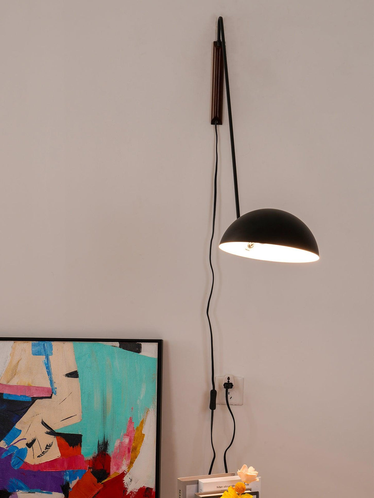 A black Clérachic wall light hangs low on a bent arm, its dome glowing, with the cord dropping to a socket by a canvas.