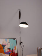 Lit black Clérachic dome wall light pointed down, with a long cord running past a leaning abstract print to a socket.