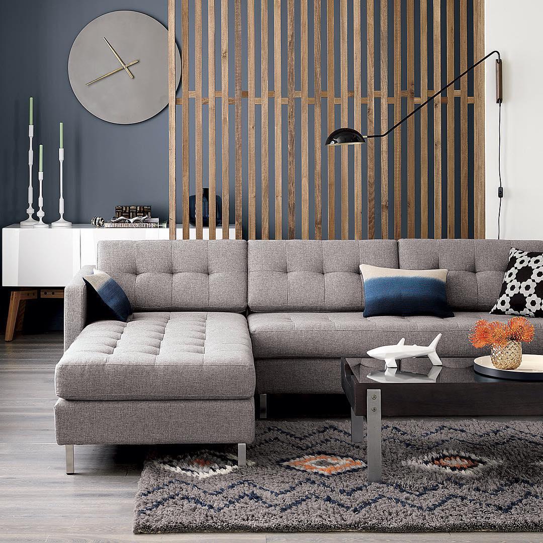 Black Clérachic wall light on a long arm beside a slatted wood divider, above a grey sectional sofa and a shaggy rug.