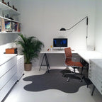 A black Clérachic wall light above a white corner desk, with a leafy palm, a tan chair and a grey wavy rug in the room.
