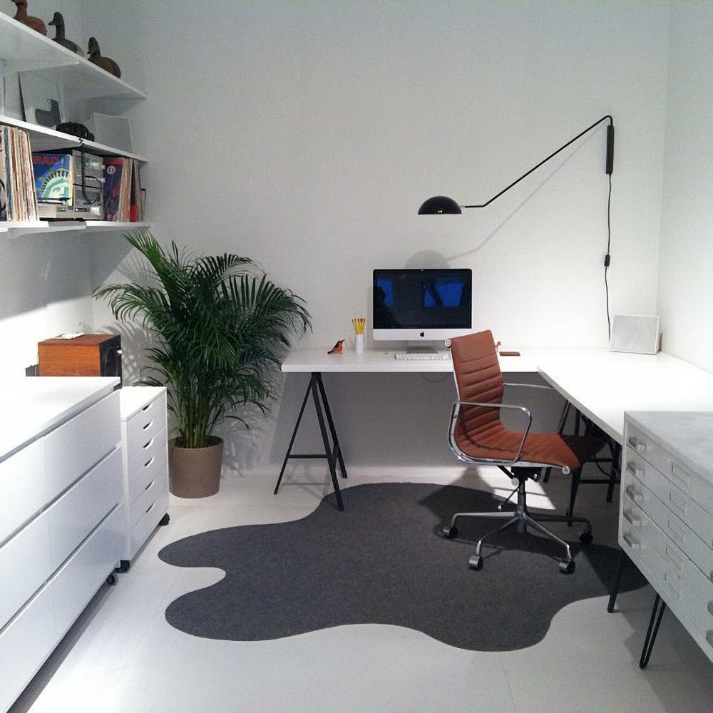 A black Clérachic wall light above a white corner desk, with a leafy palm, a tan chair and a grey wavy rug in the room.