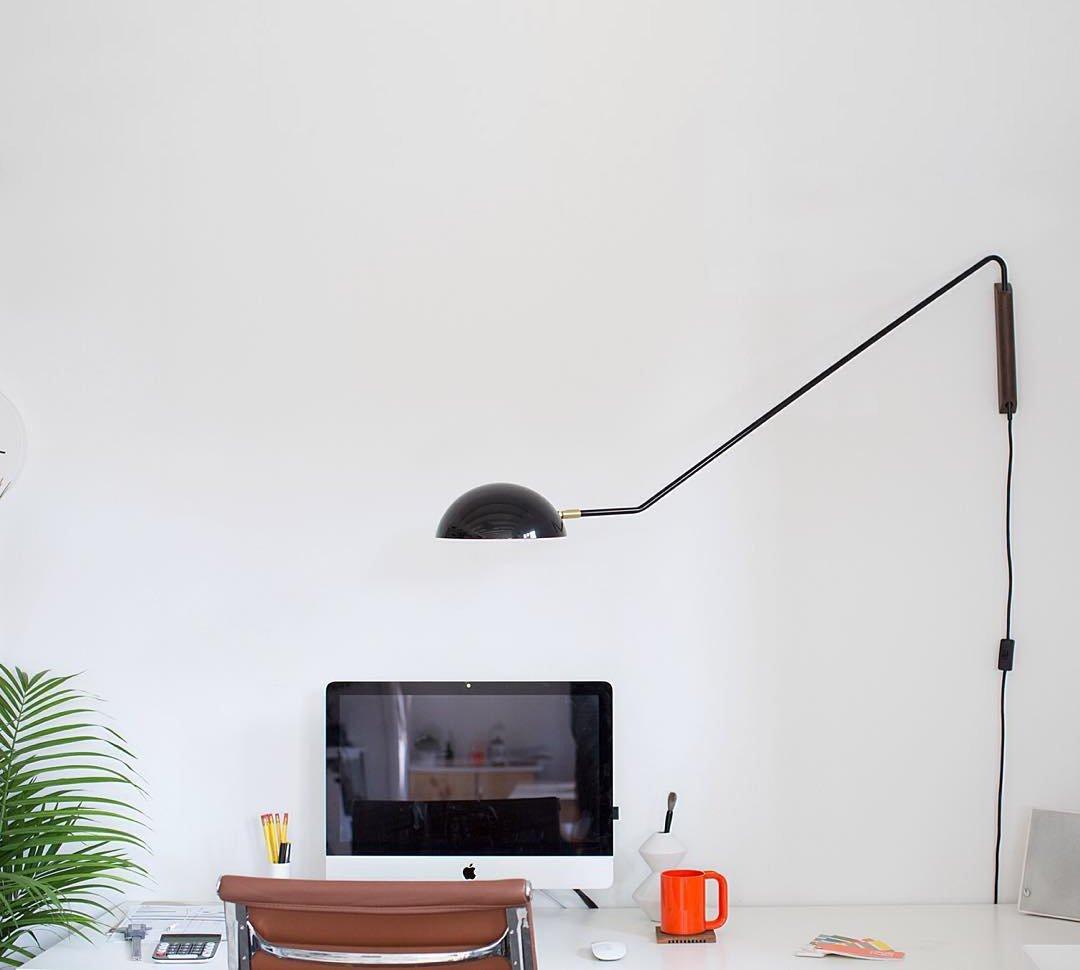 Black Clérachic swing-arm wall light on a white wall above a desk with a monitor, an orange mug and a palm leaf.