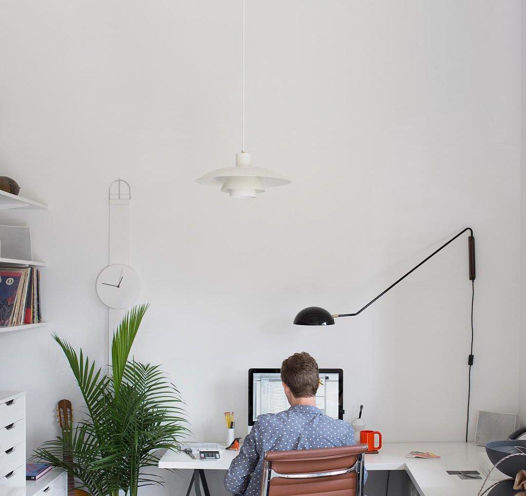 A black Clérachic wall light reaches over a home office, where someone works at a desk beside a palm and a white pendant.