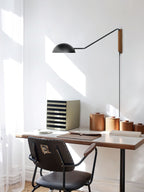 Black Clérachic wall light on a pivoting arm above a small wooden desk, with sunlight across a white wall and a dark chair.