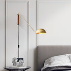 A gold and walnut Clérachic wall light above a grey headboard, on a panelled wall with a black cord and in-line switch.