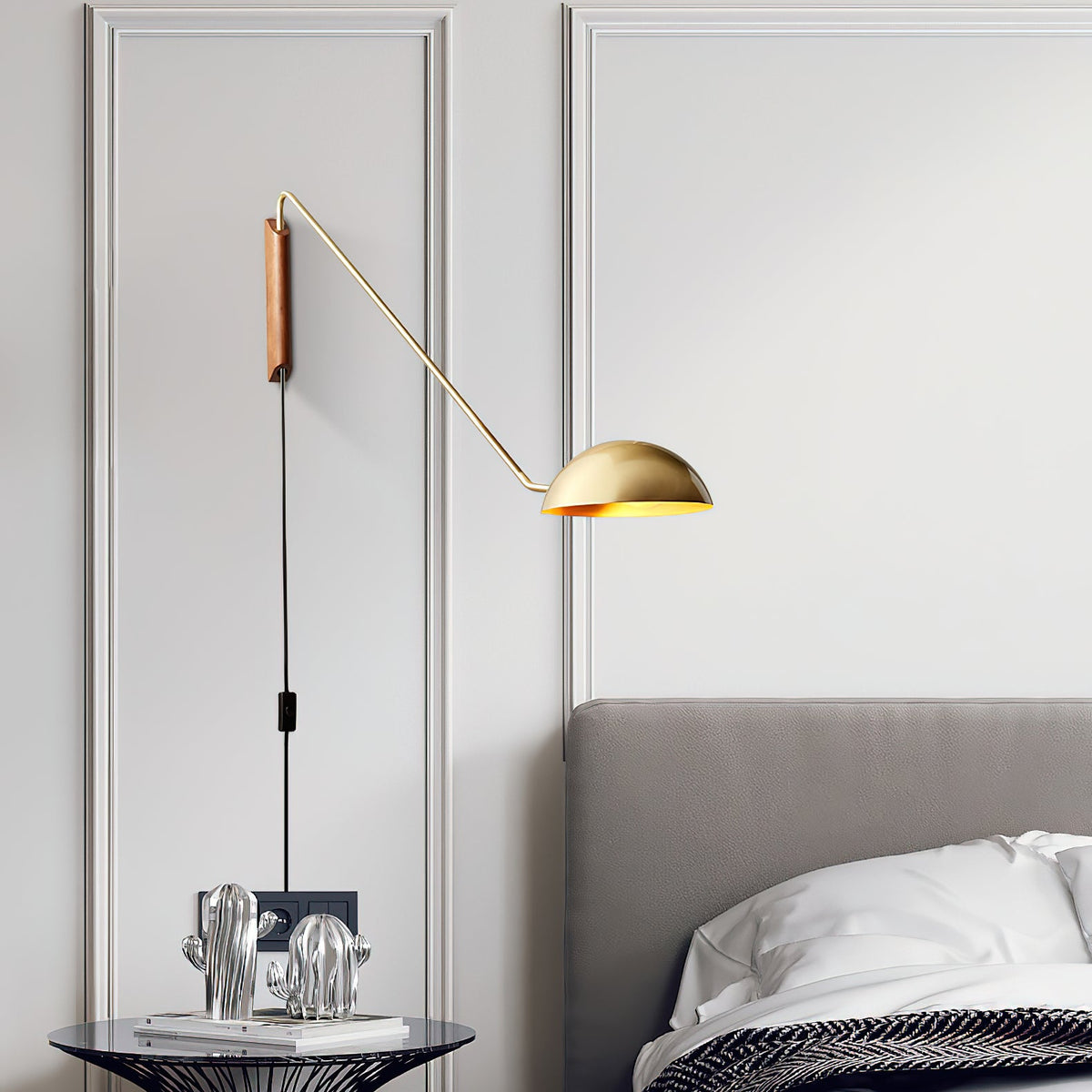 A gold and walnut Clérachic wall light above a grey headboard, on a panelled wall with a black cord and in-line switch.