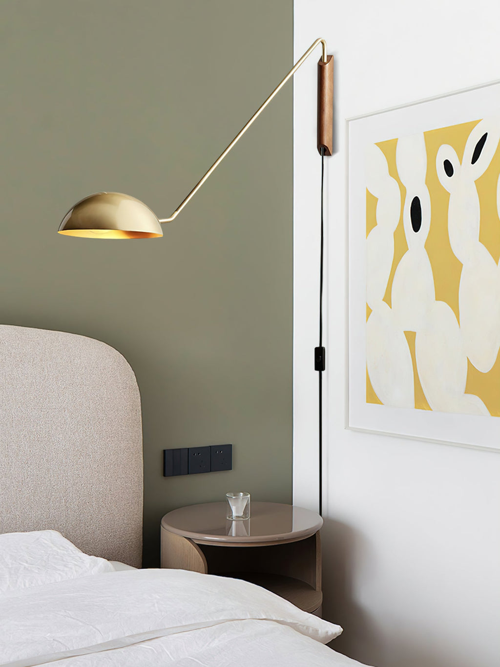 Gold and walnut Clérachic wall light on a sage green wall beside a bed, with a yellow and white abstract print nearby.